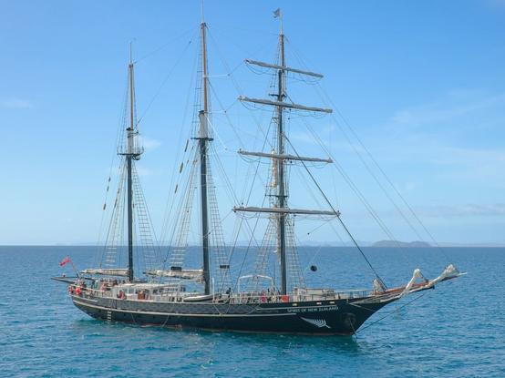 A 3 mast tall ship with its sails down, at anchor. The ship sits on calm flat water, with land visible on the horizon some distance behind it