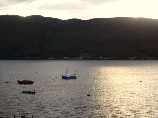 Barcos pesqueros en una ría durante la puesta de sol. Al fondo, montañas.