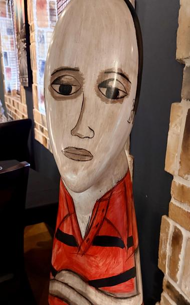 A large ceramic figurine, looking like a 3d oil painting of a sad looking balding man wearing a red shirt with their arms crossed.