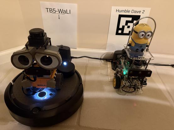 Two robots - Turtlebot 5 WaLI has a WALL-E character atop a Create3 robot base, and next to him is Humble Dave 2 which has a Minion Dave character atop a GoPiGo3 robot base.  Each robot is standing in front of their charging dock during their play time.