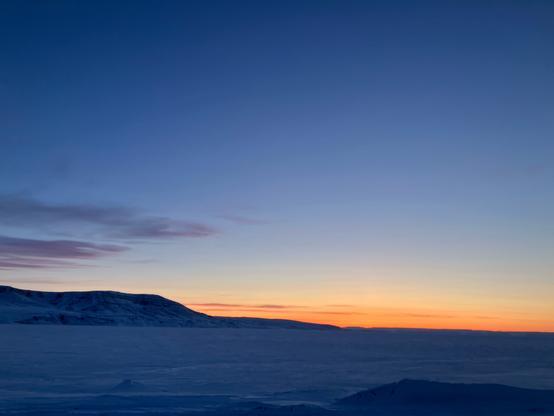 Looking across a frozen bay on the Arctic Ocean toward mountains and low hills. A deep blue sky punctuated with an orange horizon and wispy purple clouds.