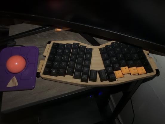 A tiny bedside table holding a monitor, a colorful 3d printed trackball, and a CNCed bamboo keyboard. It looks like a really tight fit, everything is really crammed on there. A normal mouse or keyboard, let alone both, would clearly not work in this space.
