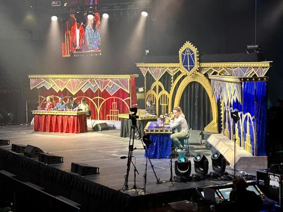 Picture of the cast of Critical Role sitting at tables on stage, from an oblique angle. The set is elaborately decorated in red, blue, and gold.