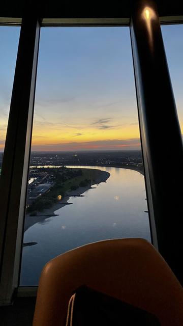 Ein atemberaubender Blick vom Düsseldorfer Fernsehturm auf eine Stadtlandschaft bei Sonnenuntergang. Die untergehende Sonne taucht den Himmel in warme Orange- und Rottöne, während sich der Rhein durch die Stadt schlängelt. Im Vordergrund ist ein Teil eines Stuhls zu sehen, der den Betrachter in die friedliche Szenerie einlädt.