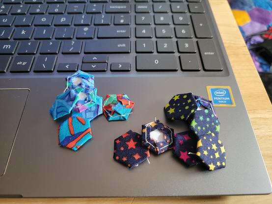 Photograph of two small piles of patchwork hexagons sitting on the corner of a laptop beside the trackpad. The hexagons have sides half an inch long, and are all sewn around paper templates. The left pile is made from scraps of a lovely Kaffe Fasset fabic in turquoise, bright red, dark blue, green and purple, and the right pile is made from fabric with a very dark blue background and a repeating pattern of tiny stars of different sizes coloured with a rainbow ombre.