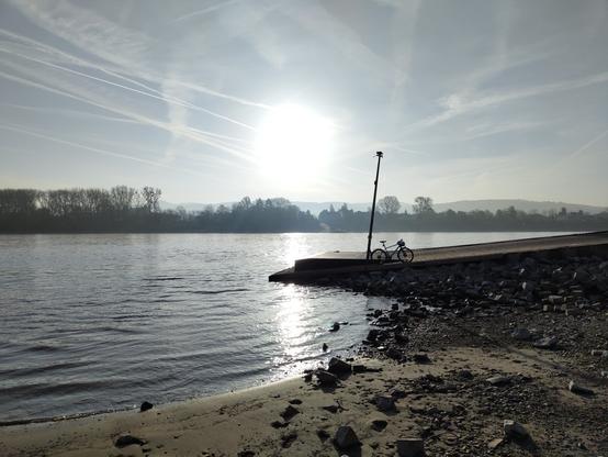 Die aufgehende Sonne am Rheinufer in Rolandseck. Der Fotograf steht im trocken gefallenen Flussbett, man erkennt den Anleger der Fähre auf dem ein Fahrrad steht.