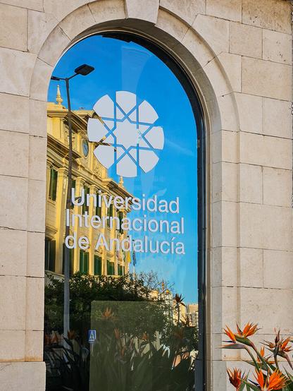 An arched window with the reflection of a large building in mainly yellow colours, with green shutters and a clock. The window glass has a logo and the words "Universidad Internacional de Andalucía".