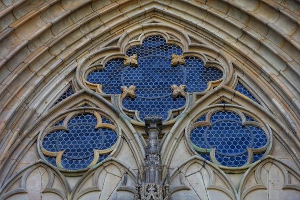 A detailed close-up captures the intricate stonework and stained glass of a Gothic archway, likely part of a cathedral. The arch is constructed from light-coloured stone, showing signs of weathering and age. The stonework features multiple layers of pointed arches, adorned with decorative mouldings and carvings.
The stained glass windows within the arch are a deep blue, patterned with a delicate, repeating geometric design of small circles. The windows are framed by ornate stone tracery, forming clover-like and quatrefoil shapes. Small, stylised floral or foliate carvings are integrated into the stone framework around the central and upper windows.
A slender, intricately carved stone pillar or mullion divides the lower section of the archway. The overall impression is one of impressive craftsmanship and architectural detail, typical of Gothic design.
