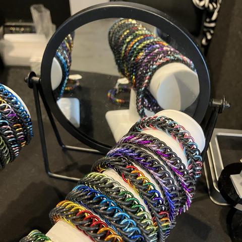 Multiple brightly coloured chainmail bracelets on a white display stand, reflected in a small round mirror.