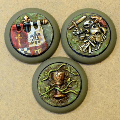 We have three flat objective markers on 32mm bases. In the top left is part of a medieval banner, in the top right is the skeleton of a rodent warrior with armour and sword and at the bottom is a golden chalice surrounded by green vines.