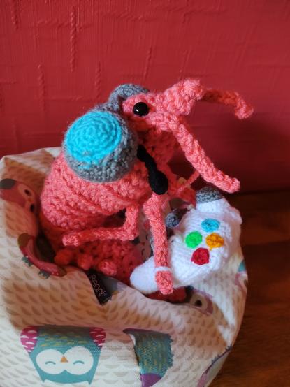 A photo of a crochet shrimp sitting on a little owl  beanbag, wearing a  grey and blue headset with a microphone and holding a white gaming controller similar to an XBox controller.
