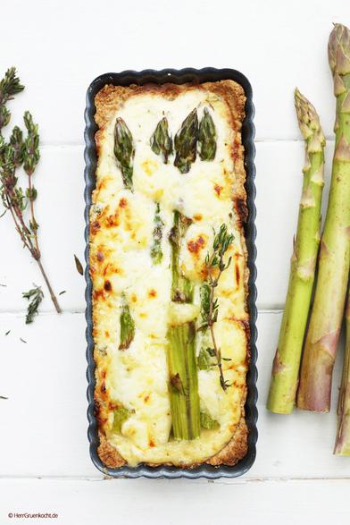 Weißer Holzuntergrund. Darauf eine rechteckige Quicheform mit einer Quiche mit grünem Spargel, in einem Bett aus Crème fraîche mit Waldhonig und Thymian
