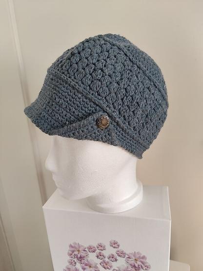 A handcrafted Newsboy hat crocheted from recycled jeans cotton yarn, displayed on a white mannequin head. The hat has a textured design with a structured brim and features an actual jeans button as an accent. The mannequin is placed on a white box with a floral design, highlighting the craftsmanship of the piece.