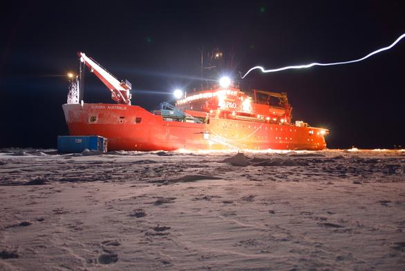 RSV Aurora Australis parked in sea ice at night, all lit up. A stream of light from the ship out to the right is somebody's headlamp