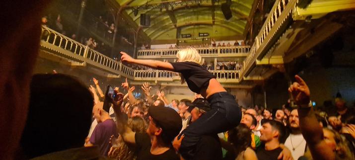 A young woman in a black tshirt and dark blue jeans on the shoulders of her partner, her left arm raised pointing to the left, in a sea of people, many doing the same, in the main room of concert venue Paradiso in Amsterdam at a sold out Mary Wallopers gig.