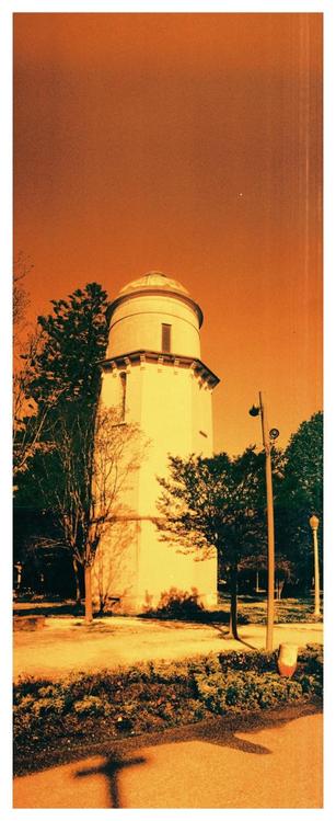 A water tower in the Parque Infante Dom Pedro in Aveiro. Taken with Harman Red redscale film the image is shades of red and orange. Above the yellow coloured tower is a ruddy orange sky, in the foreground are a pth and bushes.