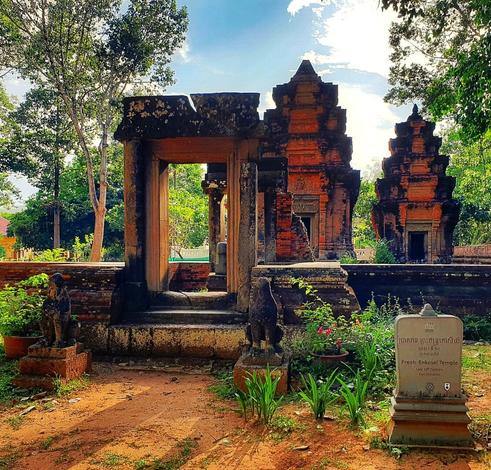Photo shows an old temple remain with two towers in later Khmer style. In front there is a gate with a small ante room and a simpler doorframe behind it. Small stairs are leading up to the main gate. On both sides of the gate is an old, low stone wall leading off to both sides. In front of the stairs there is two small guardian lion statues. There is an Apsara Marker Stone to the right, stating the name of the temple in Khmer, English, and dated to late 12th century. The track leading up to the gate is of red earth. On both sides there are flowers, plants and green turf. It is a late sunny day with lightly clouded blue sky. In the background behind the towers there are several trees to the right and left and in the distance. A couple of hours before sunset, the sunlight is warm and yellow.