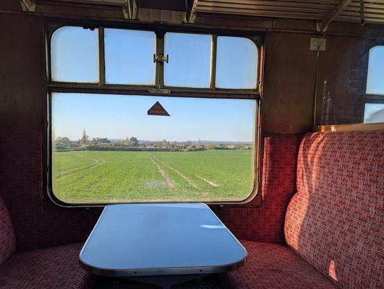 A view through the window of a vintage railway carriage of flat fields with distant trees and a church spire. Inside the carriage has wood panelling, deep plush seats in a red pattern facing a table located centrally below the window.