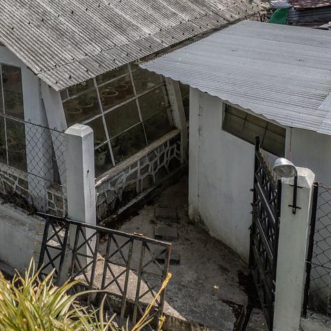 A top down photo of a greenhouse with potted plants in rows inside it visible through its glass windows. The greenhouse has a corrugated metal roof. There is a two door gate that leads toward the greenhouse where one of the doors is broken and laid down horizontally. Both the doors are made of metal and are painted black.