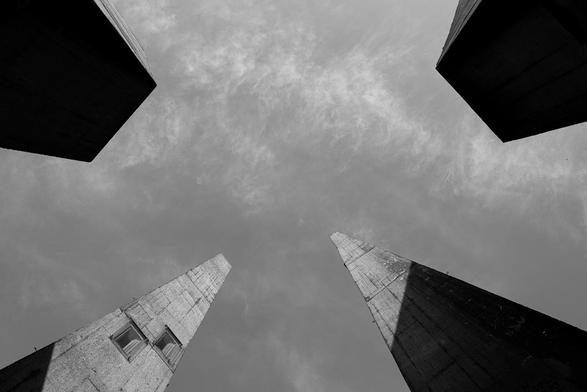 The black and white photo shows four tall, slender, rectangular concrete structures rising up against the sky from a frog's-eye view. The sky is cloudy but bright, and the concrete structures appear robust and monumental. The image appears abstract and minimalist, with the geometric shapes and the strong contrasts between light and shadow taking center stage.