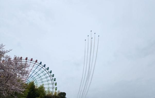 Six jet planes fly in tight formation through a cloudy sky, leaving long trails of white smoke behind them. Below, the colorful structure of a large Ferris wheel is partially visible on the left, framed by blooming cherry blossom trees and lush greenery.