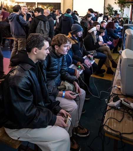 Photo qui représente une dizaine de joueurs de jeux vidéos dans une grande salle durant une convention retrogaming tous assis sur des bancs devant des anciens téléviseurs cathodique en train de jouer à diverses consoles retro.