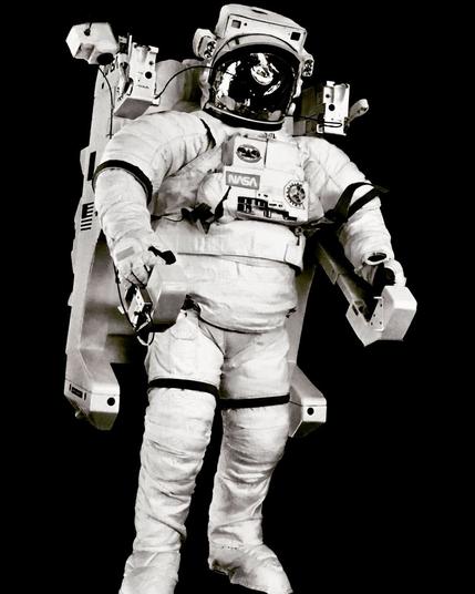 NASA astronaut wearing a white space suit with a large backpack-style propulsion unit—known as a Manned Maneuvering Unit (MMU)—against a solid black background. The suit has control panels on the chest, hand grips on extended arms, and visible NASA and American flag patches. The image is black and white.
