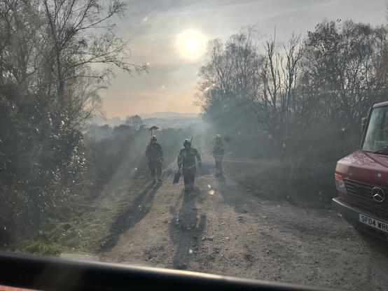 Firefighters return along a farm track to their appliance. The orb of the setting sun through heavy smoke haze behind them, long shadows cast towards the viewer. Trees to either side frame the scene. The bonnet of another appliance enters the image to the right of frame.