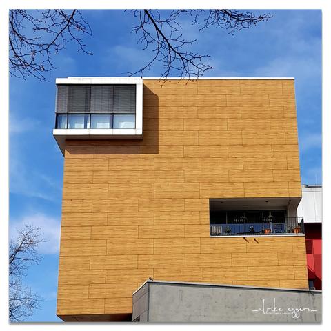 Quadratisches Farbfoto. Der obere Teil eines quadratischen Hauses mit flachem Dach, die Fassade ist mit hellem, kieferfarbenen Holz verkleidet. Es hat nur zwei große, rechteckige Fenster. Das eine ist an der rechten Seite weit unten und etwas zurück nach innen versetzt, dadurch bildet es einen Balkon, der mit einer gläsernen Balustrade gesichert ist. Das zweite Fenster ist links ganz oben und nach außen gesetzt, hat aber keinen Balkon.
Im Hintergrund ist blauer Himmel, an der linken Bildseite ragen ein paar kahle Äste ins Bild.