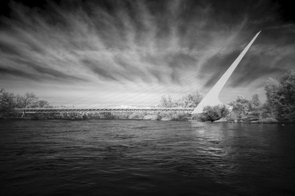 While floating the Sacramento River with my son, I had to photograph the iconic Sundial Bridge in Redding, CA. Since it’s so often photographed, I also shot it with my infrared gear. This 830nm black-and-white image reveals deep black skies and bright white clouds and foliage—well beyond the visible spectrum. The high clouds (likely contrails) made a nice sky for this dramatic capture.

Designed by Spanish architect Santiago Calatrava and completed in 2004, the 700-foot pedestrian bridge spans the river without touching it, protecting salmon habitat. Its 217-foot inclined pylon supports a cable-stay design and functions as the gnomon of a working sundial, accurate one day per year. The glass deck minimizes environmental impact and looks sleek. The bridge resembles Calatrava’s Puente del Alamillo in Spain, earning it the nickname “sister bridge” and highlighting Calatrava’s blend of art, engineering, and ecology.