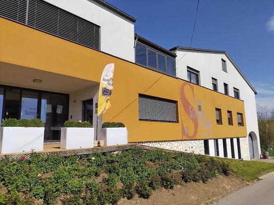 Blick von außen auf unser Weingut bei sonnigem Wetter. Davor weht eine Beachflag mit der Aufschrift "Wir haben geöffnet"