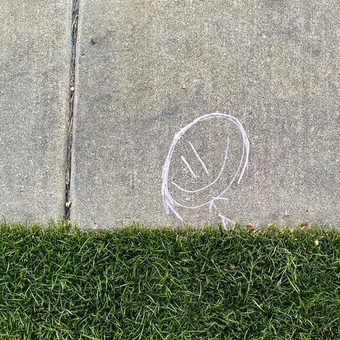 A simple chalk art stick figure with a very large head and smiling face drawn on the edge of a sidewalk to appear as though they are running through grass.
