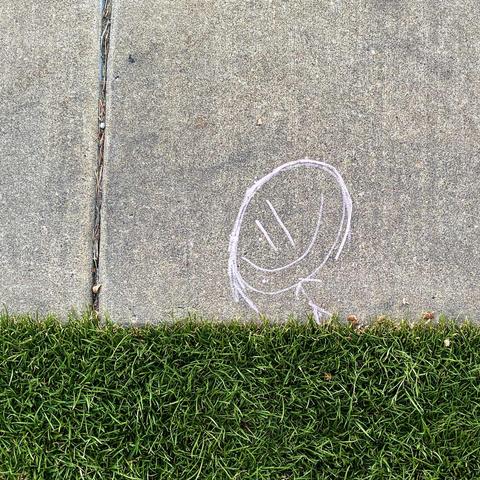 A simple chalk art stick figure with a very large head and smiling face drawn on the edge of a sidewalk to appear as though they are running through grass.