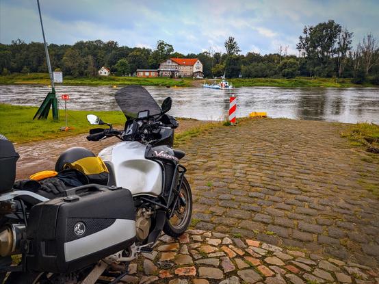 a motorbike parks at a river bench, across the river is a ferry