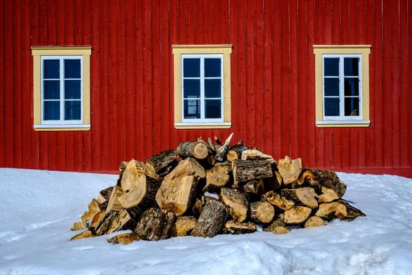 A pile of firewood in the snow. Behind the red painted wood plank siding and the 3 windows in stark contrast.