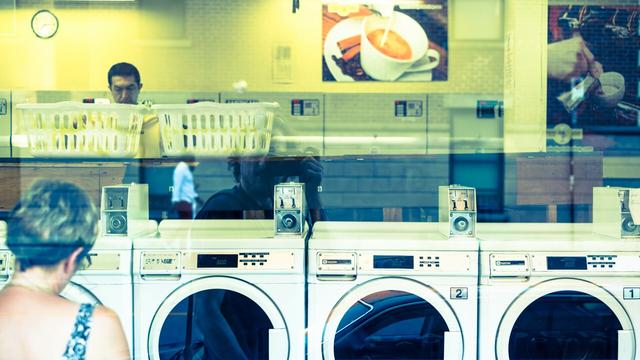 In a laundromat, a woman is using the leftmost of 4 washers. In the back a man is standing behind laundry basket. The reflection of the outside street is visible in the window the picture is taken through, including the reflection of the photographer creating layered image.