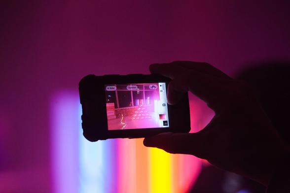 An iPhone taking a picture held by a hand, in the background light in a rainbow gradient. The iPhone show us what we can't see.