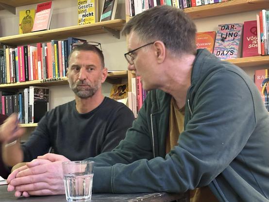 A picture of two persons seated behind a table, Tadzio Müller and myself. We're both late middle-aged persons with short hair wearing casual clothes. As I am looking toward him in conversation, Tadzio is gesturing with one of his hands while speaking. Foto taken by Suza Firyn.