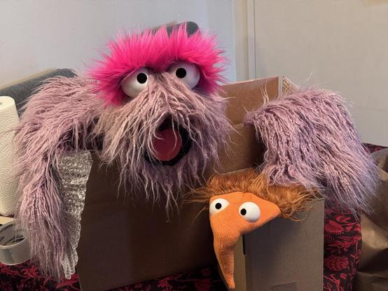 Two puppets being boxed for shipping, one a pink furry monster and the other an orange and brown Sonk puppet with a long nose
