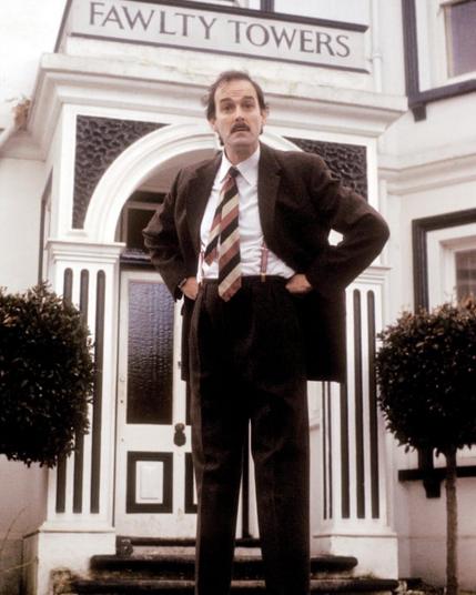 A photo of John Cleese as Basil Fawlty, standing in front of Fawlty Towers. He's wearing his trademark brown suit and tie and has his hands on his hips, with an impatient expression on his face.