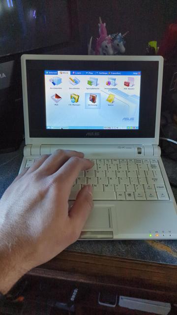 An Asus Eee PC 701, sitting on a desk. It has booted into the original Xandros OS, and seems to work well. My hand is in the frame for a size comparison; it's a very small netbook for most people with adult-sized hands.