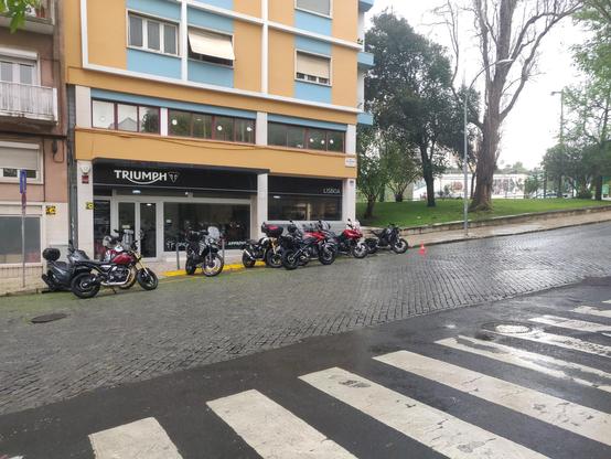 The image depicts a street scene in front of a Triumph motorcycle dealership in Lisbon. The building is a multi-story structure with a yellow and blue facade. The ground floor features large glass windows and a black awning with the Triumph logo prominently displayed. Several motorcycles are parked in front of the dealership, including models in black, red, and silver. The street is paved with cobblestones, and a pedestrian crossing is visible in the foreground. A traffic cone is placed near the motorcycles, and a tree-lined park area is visible in the background. The sky appears overcast, suggesting a cloudy day.