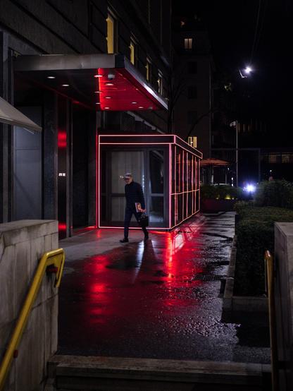 A man walks past a closed restaurant "dehor" at night after the rain. The dehor is illuminated by red LED strips and the rest of the scene is otherwise quite dark.