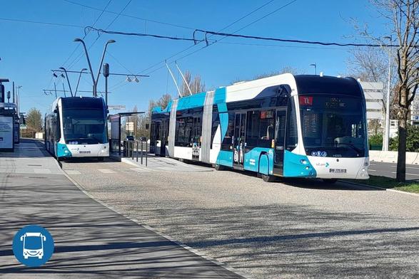 The image shows two modern trolleybuses at a stop under a clear blue sky. The trolleybuses have a sleek design, primarily white with light blue accents and dark tinted windows. The one on the left is stationed at the platform, while the one on the right appears to be about to depart, as suggested by its position. Overhead, electric wires are visible, connecting to the buses, which are articulated to navigate city streets. The bus station is minimalistic, featuring glass panels, with a few bare trees in the background. A blue circular logo with a bus icon is visible in the bottom left corner.