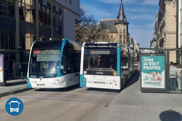 The image depicts an urban street scene with two buses and a bus stop. On the left, a blue and white bus is parked, showcasing a modern, streamlined design. To its right is another similar bus, slightly larger and partially obscured by the first. Both display the "HESS" logo. To the right of the buses, a billboard advert with a graphic of a dog and text is mounted inside a glass shelter, where several people are waiting. In the background, the street is lined with historic buildings featuring ornate architecture, and a tall, pointed tower rises prominently in the distance beneath a clear blue sky.