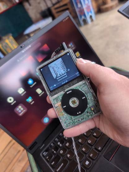 A modded transparent iPod Classic 5th Gen running a GNOME themed RockBox playing "Generational" by "408". An out of focus ThinkPad running Fedora Silverblue with GNOME desktop in the background.