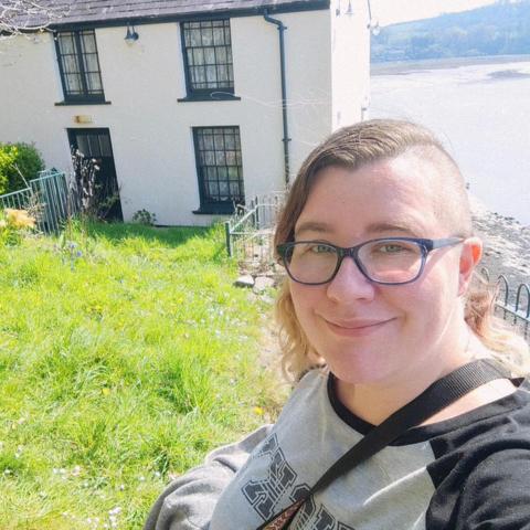 A smiling white woman in her late 30s is looking at the camera. She's got short hair on one side of her head and long brown hair on the other. She's wearing blue glasses and a grey and black top that reads "lucky" backwards (since the photo is mirrored by the selfie camera). In the background is a cream coloured building, green grass, and a view of the sea.
