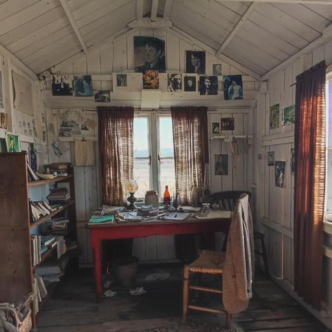 An interior view of a shed set up as a writing workspace from the past: unframed photos adorn the white walls, see through curtains in browny-orange hang in the windows, and there's a stunning view of the sea. A cluttered bookcase full of fallen over books can be seen to the left, while the middle is taken over by a table and a chair with a cardigan on it. The table is covered in notes, half written book pages, writing implements, books, and the debris of creativity or madness. (This is where Dylan Thomas wrote a bunch of well known stuff.)