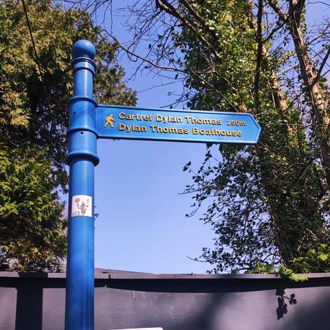 A blue metal sign directing you to Dylan Thomas' boathouse in both Welsh and English.