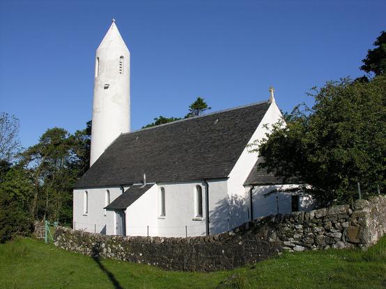 Kilmore Church at Dervaig. The image shows a rear quarter view of a white church beyond a grey stone wall. At the far end of the church is a white circular pencil-like tower. The sky is blue.